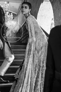 An ivory front open tissue shirt with heavy embroidery decorated with dull golden and champagne kora, dabka, tilla, naqshi, sequins and swarovski. It is further allured with cutwork and floral embroidery. Its umbrella sleeves is embellished with delicate zari work. Its black inner shirt is also embroidered. This outfit is artistically coordinated with black chiffon sharara which is allured with floral embroidery. This outfit is fully scalloped and enhanced by a thick floral embellished pattern. Dress comes with ivory dupatta which has finished edges and having sprinkled sequins pattern all over it.