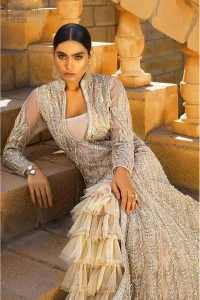 A fusion of traditional and latest fashion making the outfit trendy and unique. This angrakha style floor length maxi is heavily hand crafted with floral embroidery on the bodice, floral booties and vertical lines all over the dress. Back is also enhanced with vertically designed floral booties and swarovski crystals lines all over. The embroidery is done in the shades of silver and dull golden zardozi work. Style it up with frilled lehenga to give it a regal look. This outfit is comprises with fawn net lehenga sprinkled with sequins spray all over.