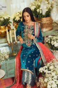This dazzling yet elegant ensemble is perfect to make a statement at any event.  Long shirt for party dress in teal blue colour is embellished with multiple colour embroidery which is prominent with dabka, naqshi, stones, tilla, crystals work. The boat shape neckline is decorated with embroidery and with full-length sleeves. The fabric used for the shirt is velvet. It is paired with jamawar trousers in the same colour that is unique in style. Finish this party wear with a scalloped dupatta framed with a four-sided border and ting floral motifs all over that is the perfect choice for a party.