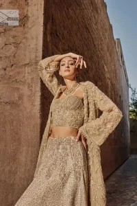 This modern piece is all about extraordinary details and aesthetic vibes! A beige organza blouse is heavily embellished with dabka, naqshi, tilla, Resham, dabka, sequin and Kundan. This gown blouse dress has Mughal-inspired motifs, geometrical patterns and floral motifs. The blouse has a sweetheart neckline or a more upscale appearance and feel. Further, the gown has full sleeves adorned with floral patterns. It is supported by a flared lehenga with an embellished border for additional comfort.